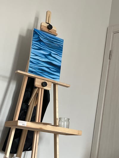 The image shows an easel holding a painting of abstract blue wavy lines. The easel is made of light-colored wood and has a black bag folded beneath it. A clear glass jar is attached to the top of the easel. The painting is on a flat surface supported by the easel. In the background, there is a section of a wall with a light-colored cabinet door at the top, suggesting an indoor setting. The lighting is even, indicating an indoor environment without direct sunlight. There is no discernible text in the image, except for what appears to be a small label on the easel. The scene appears to be set up for artistic creation or display.