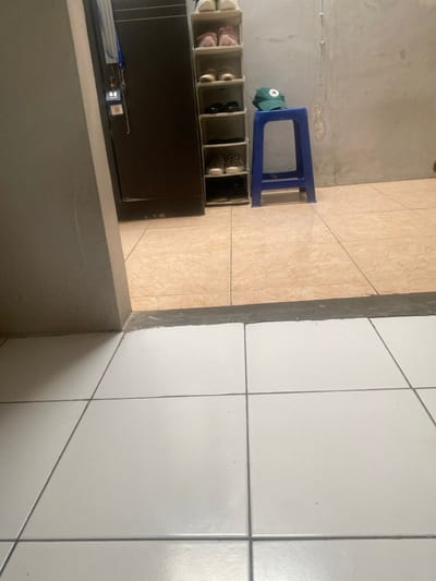The image depicts an interior scene, observed from a low vantage point across a transition between two distinct floor areas, located in Cimenyan, Indonesia. In the foreground, the floor consists of large square white ceramic tiles with dark grey grout lines. A dark grey or black concrete threshold separates this area from the room beyond.
The room beyond the threshold features light brown/beige ceramic floor tiles. To the left, a dark brown, vertically oriented structure, possibly a cabinet or doorframe, is partially visible. Attached to its left side are a blue lanyard and a small white rectangular object displaying what appears to be a QR code or barcode. Adjacent to the right of this structure stands a narrow, tall shoe rack with six open shelves. From top to bottom, the shelves hold: two pairs of light-colored athletic shoes, two pairs of pink athletic shoes, two pairs of light brown/beige shoes, two pairs of black closed-toe shoes, two pairs of checkered pattern slip-on shoes, and a single dark shoe on the bottom shelf.
To the right of the shoe rack, a solid blue plastic four-legged stool is positioned against a plain, light grey concrete or stucco wall. A green baseball cap with a white star logo is resting on the seat of the blue stool. The overall environment appears well-lit by ambient light. No individuals or active interactions are present in the scene.