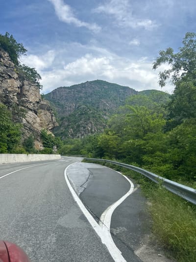 The image captures a road winding through a mountainous landscape, likely near Krichim, Bulgaria. The asphalt road has a white line marking the edge, and a metal guardrail runs along the side of the road. On the other side of the road is a rocky cliff face covered in trees. A mountain rises in the background, partially obscured by trees. The sky above is cloudy. A small section of a red vehicle is visible in the lower right corner of the image.