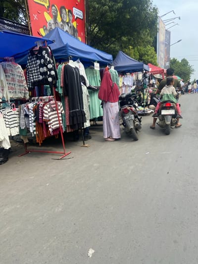 The image shows a street market in Margaasih, Indonesia (latitude: -6.953, longitude: 107.564). Several vendors have set up shop along the side of the street, displaying clothing items on racks. There are several customers browsing through the wares. One woman is standing in front of a clothing rack, wearing a long, pink hijab, examining the items. A man and a young girl are standing on the side of the street, looking at the merchandise. The man is wearing a red shirt and a backpack. The girl is wearing a pink dress. Behind them is a motorbike. The street is paved with asphalt. The sun is shining and the sky is cloudy. There are trees and buildings in the background.