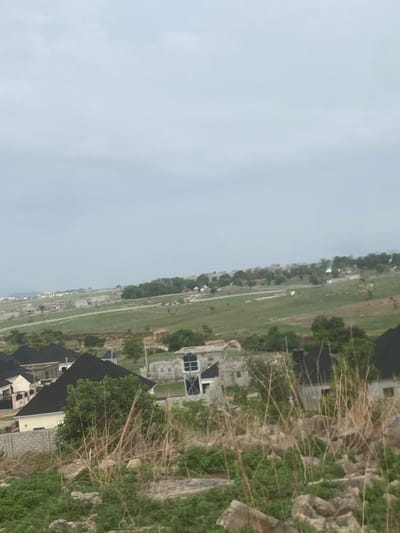 The image shows an elevated view in Iddo Tudunwada, Nigeria, looking down towards a flat expanse. There's a visible contrast between undeveloped land, with sparse vegetation and rocky terrain in the foreground, and the development of houses with dark roofs. A long straight road runs through the middle of the scene, with buildings and occasional trees lining its sides. The sky is overcast.