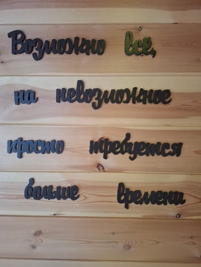 The image shows a wooden wall with several phrases written in Russian, made of black, cut-out letters, possibly wood or plastic. The phrases are arranged on separate wooden planks.
From top to bottom, left to right, the phrases are:
- "Возможно" (Vozmozhno - Possibly)
- "все" (vse - everything) - this word is green with a textured surface.
- "на" (na - on)
- "невозможное" (nevozmozhnoye - impossible)
- "просто" (prosto - simply)
- "требуется" (trebuyetsya - requires)
- "больше" (bol'she - more)
- "времени" (vremeni - time)
The wall appears to be made of light-colored wooden planks, possibly pine, with visible grain and knots. The lighting suggests an indoor environment, possibly a room or a covered outdoor area. The overall aesthetic is rustic and decorative.
There are no people or animals visible in the image. The scene is static and appears to be a close-up shot of the wall art. The context of Sukhoy Ruchey, Russia is not visually evident from the image itself.