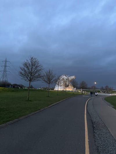 The scene depicts The Kelpies, two large horse-head sculptures, illuminated at dusk or early night in The Helix park, Falkirk, United Kingdom. The sky is heavily overcast with dark grey-blue clouds.
In the foreground, an asphalt pathway with a distinct yellow dividing line runs from the bottom center, curving towards the right. To its right, a narrower path with a gravel edge is visible. To the left of the main asphalt path is a curb, followed by a strip of neatly mown green grass.
The midground features a wider expanse of green grass on the left. Several bare-branched deciduous trees are sparsely scattered across this grassy area, extending towards the background. The Kelpies sculptures, composed of metallic segments, are prominent on the right, bathed in bright, warm upward-facing illumination. In front of the sculptures and to the right of the asphalt path, a group of four individuals is walking away from the viewer. They appear to be adults; one pair leads, followed closely by another pair. Streetlights are visible along the pathways and near the sculptures, emitting a yellowish light.
In the far left background, a large electricity transmission tower (pylon) stands against the dark sky. The distant background beyond the sculptures shows indistinct dark land features and sparse vegetation.
