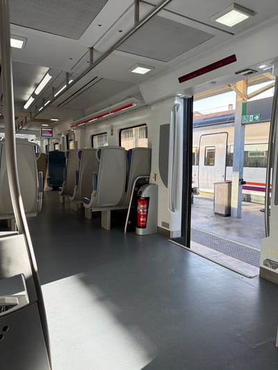 The image shows the interior of an empty train carriage, with a view through an open doorway to a train on an adjacent platform. The train carriage is modern and clean, with grey seats arranged in rows and a grey floor. There is a fire extinguisher mounted on the wall near the open doorway. Outside the train, a station platform is visible, with a silver trash bin and a sign indicating accessibility and an exit. The train on the platform is white with a purple and orange stripe. The lighting suggests it is daytime, with sunlight entering the carriage and casting shadows on the floor. Based on the red sign displayed inside the carriage that reads "DESTINACIÓ: PORTBOU," the location is likely a train station in or near Figueres, Spain, as Portbou is a town on the French border accessible by train from Figueres.
