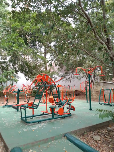 The image shows an outdoor fitness park in Cuiabá, Brazil, with various exercise machines situated on a green rubberized surface. The machines are predominantly orange and green, with some metal components. In the foreground, several pieces of equipment are visible, including what appears to be a leg press machine and an elliptical trainer. In the background, more fitness equipment is scattered throughout the park, with trees and foliage providing shade. The ground is covered with a light green surface, and fallen leaves are present on the ground around the park. The overall impression is of a public space designed for outdoor exercise and recreation.