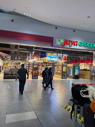 A wide shot captures the interior of a grocery store, likely Jaya Grocer in Malaysia, with a focus on the entrance and a section of the aisle. The store's signage, "Jaya Grocer," is prominently displayed in bright red and green letters. Inside, shelves are stocked with various products, including what appears to be food items and souvenirs.

Several people are visible in the scene. In the foreground, a man wearing a dark t-shirt and black pants walks away from the camera, his back to the viewer. Further into the store, a group of three people, two women and possibly a man, are walking together, their backs also to the camera. One of the women is wearing a patterned dress, and the other is in dark clothing. The overall lighting suggests it is daytime, as the interior is well-lit, and there are no indications of natural light from windows. The floor is tiled with large gray tiles.

Prominently displayed on the shelves are items with text such as "Malaysian," and on a digital screen, "10% Off." Other visible text includes "EMPIRE SUSHI." The overall atmosphere appears to be one of a busy shopping environment.