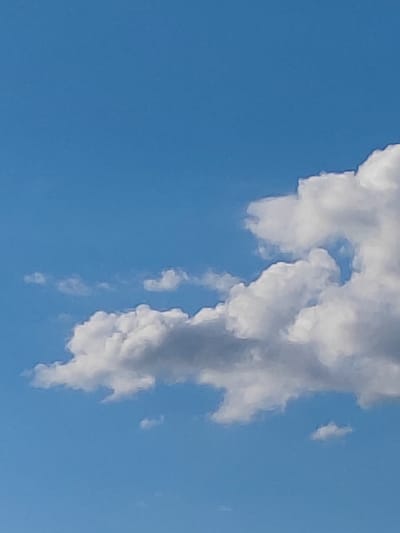 This is a vertical, close-up shot of a blue sky with scattered cumulus clouds. The clouds are mostly white and fluffy, with some darker gray shadows indicating depth and texture. The sunlight suggests it's daytime. The overall impression is peaceful and serene. There are no people, objects, or text visible in the image. The setting is outdoors, in a clear sky.