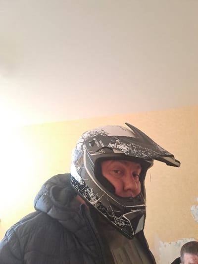 The image displays an interior scene, identified as being captured in Bolshaya Elhovka, Russia. The primary subject, an adult male, is positioned in the foreground, facing directly towards the viewer. He is wearing a dark grey and white full-face off-road motorcycle helmet, emblazoned with a "hot traxx" logo and a distressed, shattered-effect graphic design. His face, with light skin and a neutral expression, is visible through the helmet's open eye port. He is dressed in a dark, possibly black, hooded puffer jacket over a lighter-colored shirt. A second individual's head is partially visible out of frame in the bottom right corner. The background features two distinct interior walls: the left wall is a uniform light yellow or beige, while the right wall shows significant patches of peeled plaster or paint, revealing an underlying lighter surface. The ceiling is white and appears evenly lit. No direct interaction between the two individuals is discernable within the visible frame.