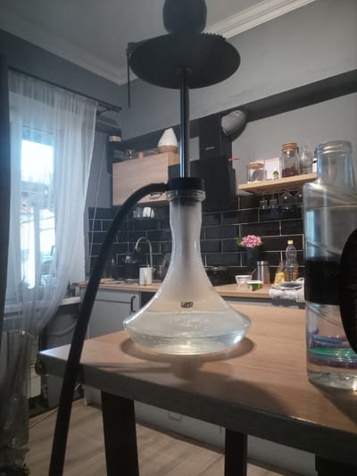 A hookah sits on a wooden table in what appears to be a kitchen or dining area. The hookah has a clear glass base filled with water, a metal stem, and a black hose. Behind the hookah, a kitchen sink is visible with black tiled backsplash. A window with sheer curtains is on the left side of the frame. On a shelf above the sink are several glass jars and a small diffuser. To the right of the hookah, a large glass bottle with colored rings inside is also on the table. The overall impression is of an indoor scene, likely a residential setting. The location in Nalchik, Russia, is not visually evident from the image itself.