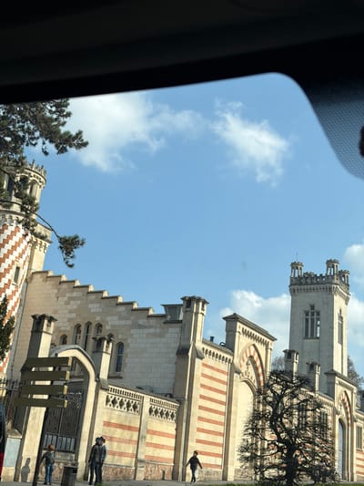 An outdoor daytime image, captured from inside a vehicle, depicts a section of the Villa Demoiselle in Reims, France, under a clear blue sky with scattered white clouds. The prominent architectural complex, a blend of Art Nouveau and Neo-Gothic styles, features facades constructed with light-colored stone and distinctive horizontal bands of red brick masonry. On the far left, a circular, crenellated tower displays a red and white chevron pattern. Adjacent to it, the main structure exhibits a battlemented roofline and arched windows. A dark, bare deciduous tree is positioned in the mid-ground on the right, partially obscuring a longer wall section of the building, which also features the alternating red brick and light stone pattern with arched openings. A tall, rectangular crenellated tower with elongated arched windows rises prominently on the far right. On the paved sidewalk in the foreground, five individuals are present: three adults and two children. Near a decorative metal gate and a multi-paneled signpost on the left, an adult in a light blue top and a child in a dark jacket stand adjacent to a dark trash receptacle. Further right, another adult in dark clothing and a child are partially visible near an arched entrance. A third child, wearing a dark jacket and light pants, walks towards the right side of the frame. Strong sunlight casts distinct shadows on the ground.