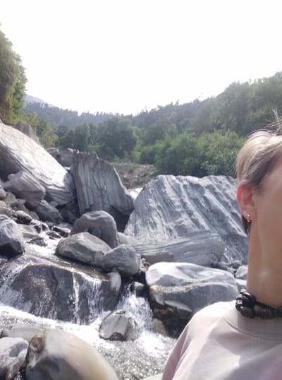 An over-the-shoulder shot captures a person's profile from the right side of the frame, facing left. The person has light-colored hair, an ear with a small gold hoop earring, and is wearing a light-colored t-shirt and a dark beaded necklace. The background is a natural landscape featuring a rocky riverbed with flowing water. Large, smooth, grey rocks are scattered throughout the scene, some of which have distinct striations. Beyond the riverbed, a lush green forested hillside rises, with the sky a bright, washed-out white at the top of the frame. The scene appears to be a tranquil outdoor location, possibly in the city of Baragran, India, given the natural elements.