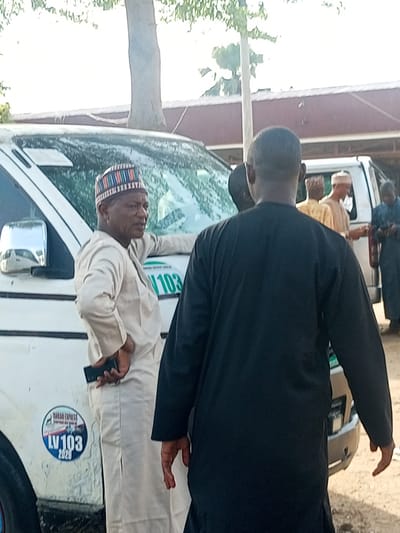The image shows a group of men standing near a white van in an outdoor setting. One man, wearing a light-colored traditional outfit and a colorful cap, leans against the van and holds a phone in his hand. The van has a logo that reads "TAKARI EXPRESS LV103 2020." Another man, wearing a black robe, stands with his back to the camera. Other men in traditional attire are visible in the background. The location is assumed to be Bauchi, Nigeria, but there are no other landmarks to confirm.