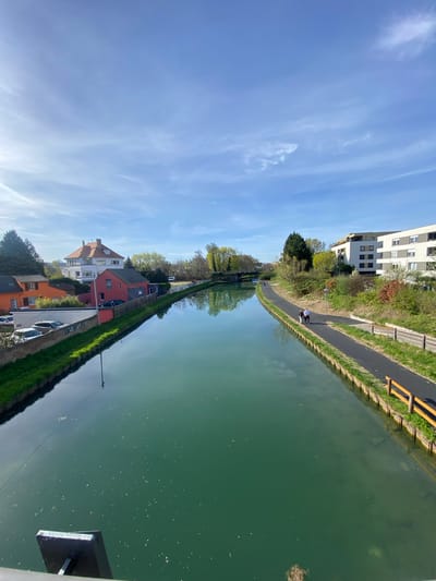 A vertical eye-level outdoor shot shows a canal in Hœnheim, France, bordered by a paved path and a grassy bank on the right and residential buildings separated by a green hedge on the left. The canal water is green and reflects the sky. Two people walk along the paved path at a distance. The sky is blue with thin clouds. A bridge rail is visible in the bottom right corner.