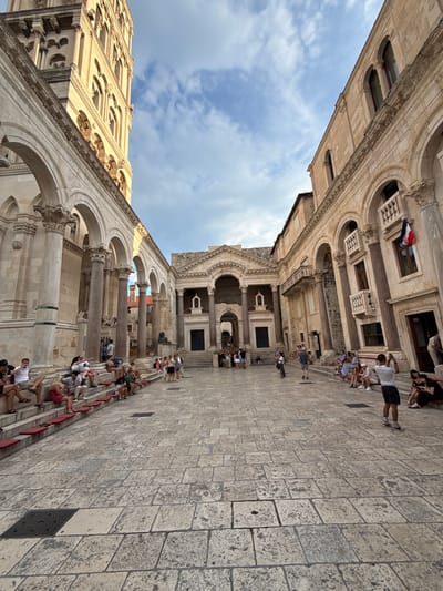 The image captures a bustling scene within the historic Diocletian's Palace in Split, Croatia. The perspective is a dramatic low-angle shot looking upwards, emphasizing the grandeur of the ancient architecture. Large stone buildings with arched windows and columns dominate the frame, bathed in natural light, suggesting it is daytime. The sky above is a mix of blue and white clouds, indicating fair weather.
Numerous people are scattered throughout the scene, many of whom are seated on stone steps, appearing to be tourists or visitors taking a break. They are dressed in casual summer clothing, with a mix of shorts, t-shirts, and dresses, consistent with warm weather. Some individuals are actively engaged in taking photographs with their phones, highlighting the tourist activity. The ground is paved with large, worn stone blocks, characteristic of ancient city squares.
Notable details include a Croatian flag flying on one of the buildings, providing a strong locational cue. The architecture itself, with its intricate carvings and imposing scale, speaks to the historical significance of the site. The overall atmosphere appears relaxed and leisurely, with people enjoying the historical ambiance and the pleasant weather. The image is a snapshot of daily life within a world-renowned heritage site, showcasing the blend of ancient history and modern tourism.