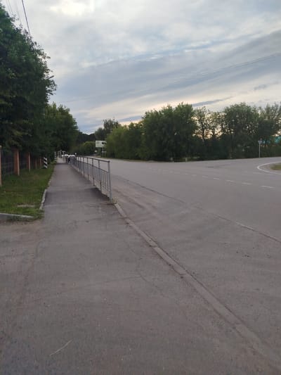 This media file depicts an outdoor scene in Zavodoukovsk, Russia, likely during the late afternoon or early evening, given the overcast sky and soft lighting. The image shows a paved road with clear lane markings and a sidewalk running alongside it. A metal railing separates the sidewalk from the road in some sections.
The setting is urban or suburban, with a dense line of green trees bordering the road and sidewalk. A fence, partially visible on the left side of the image, suggests a residential or private property area adjacent to the sidewalk. In the distance, several cars are visible on the road, indicating normal traffic flow. One person appears to be walking on the sidewalk in the distance.
The sky is cloudy, with light and dark streaks of clouds suggesting a dynamic, perhaps somewhat cool, weather. There are no strong emotional cues conveyed by the scene; it is a straightforward depiction of a common roadside environment.
There is a sign visible in the distance on the right side of the road, but the text on it is too indistinct to be read. The overall impression is one of a quiet, functional public space.