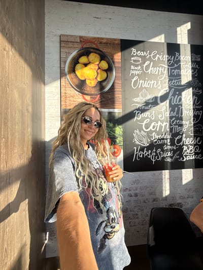 A young adult woman with long blonde hair, wearing sunglasses and a grey t-shirt with a Mickey Mouse print, smiles at the camera while holding a red drink. She is standing in front of a wall with a large menu board that lists various food items in white chalk lettering, including "Beans," "Crispy Bacon," "Cherry Tomatoes," "Onions," "Chicken," "Corn," "Cheese," and "BBQ Sauce." There is also a framed image of potatoes in a colander on the wall. The environment appears to be a restaurant, with wooden wall paneling and brickwork visible. Sunlight streams in from the side, casting shadows across the scene. The location is stated to be Шебунино, Russia.
