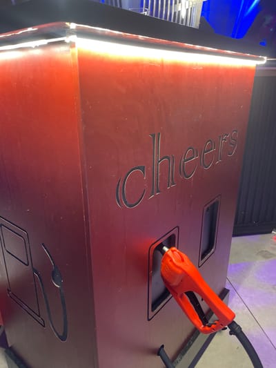 The image shows a close-up of a red, cabinet-like structure that appears to be a novelty "gas pump" for drinks or something similar. The word "Cheers" is carved into its side. An orange nozzle, resembling a fuel pump nozzle, is inserted into a rectangular opening on the front of the cabinet. The structure is illuminated from above by bright white LED lights. The background is blurred, with hints of blue lighting and what looks like a metal railing. There are no people visible in the frame. The environment appears to be indoors, possibly a bar or entertainment venue. The scene is not identifiable as being in Hadera, Israel, based on visual cues.