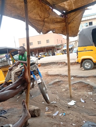 In Tudun Nupawa, Nigeria, a man wearing a green soccer jersey and khaki pants sits on a blue motorcycle parked next to a yellow auto rickshaw (tricycle taxi). The motorcycle faces toward a building with multiple windows across the street. The ground is dirt and has scattered debris. Another person's bare feet are prominently visible in the foreground. The scene is under a makeshift canopy supported by wooden poles. More rickshaws and people are visible further down the street.