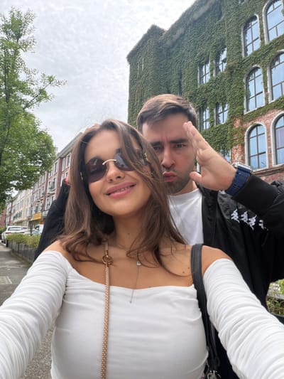 A man and a woman are posing for a selfie outdoors, likely in an urban area. The woman is in the foreground, wearing sunglasses, a white top with long, gathered sleeves, and necklaces. The man is behind her, wearing a black jacket over a white shirt and a blue watch. His hand is raised in a casual gesture. Behind them are buildings; one is covered in green ivy, and another is made of red brick. The sky is overcast. No clear indications suggest the location is Zhangjiajie, China.
