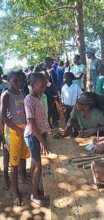 The image shows a group of children and adults gathered in an outdoor setting, likely in a village in Burundu, Kenya, located at latitude 0.529 and longitude 34.746. The children are wearing a variety of clothing, including school uniforms and casual wear. The adults are wearing casual attire. There is a wooden table in the center of the scene, with various objects on it. One adult female appears to be speaking to a child standing in front of her. Several children stand behind them, and an adult male is sitting to the right of the table. The background shows trees and foliage, with bright sunny weather. The scene appears to be a gathering of children and adults in a casual setting. There are no signs of any illegal or harmful activity in the image.