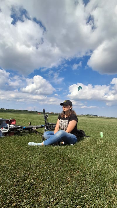 A woman with long, dark curly hair, wearing a black baseball cap with a yellow emblem, a black t-shirt with a white graphic print, and blue jeans, is sitting cross-legged on a grassy field. Her light blue socks are visible. To her left, a bicycle is lying on its side, with its wheels and handlebars partially visible. A black backpack is situated behind her. In the background, a teal kite is visible in the sky, which is filled with fluffy white clouds against a blue sky. The field appears to be an open, undeveloped area, possibly on the outskirts of the city of Kempelevo, Russia, given the sparse vegetation and distant low-rise buildings. A green can is placed on the grass to the right of the woman. The overall scene suggests a relaxed outdoor activity on a sunny day.
