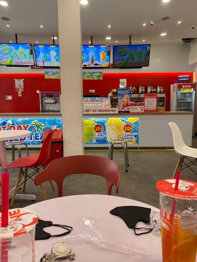 The image shows a cafe interior with a white table and a black face mask on it. Two red chairs are in front of the table. A glass of orange liquid with a red lid and a straw is in the foreground. Behind the table, there is a counter with a red-and-white sign reading "1997-ICE CREAM & TEA". On the counter are displays featuring "Kiwi Fruit Tea" for 16k and "Fresh Squeezed" for 10k. Two red chairs are facing the counter. The walls are red with advertising displays showing images of various drinks. The location is in Kecamatan Piyungan, Indonesia.