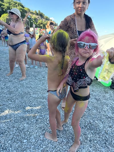 This media file captures a group of people on a pebble beach under a clear sky, suggesting a daytime setting. The participants are adorned with vibrant colored powders, indicative of a Holi festival or a similar celebratory event involving the throwing of colors.
In the foreground, a young person with bright pink hair and sunglasses is holding a packet of yellow powder. To their left, an adult wearing a leopard print cover-up and a black bikini top looks towards the camera with a neutral expression. Another young person, facing away from the camera, has green powder on their hair and back, and is wearing a striped bikini. Their legs, dusted with yellow and pink powder, are visible resting on the pebbles. Further back, other individuals are seen enjoying the event, some with visible colored powders on their clothing and skin. One person in the middle ground is wearing a patterned bikini. In the bottom left, an older woman with a straw hat and sunglasses is also in a bikini and appears to be looking down.
The background shows a lush hillside with trees, typical of a coastal landscape. The presence of a light-colored tent or canopy can be seen behind the people in the foreground, possibly providing shade. The overall mood appears to be festive and joyful, with the bright colors adding to the lively atmosphere. No discernable text is clearly visible in the media file, though there is some writing on the yellow packet being held.