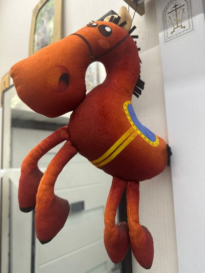 This media file features a plush toy horse with a saddle. The horse is primarily brown with black yarn for its mane. It has a blue and yellow patterned saddle and small white eyes with black pupils. The toy is positioned as if it is lying down.
In the background, the top portion of the image shows a white surface with a metallic emblem. The emblem appears to be a crest with a shield shape and intricate designs within it, including what looks like a stylized cross or figure. Below the emblem, some text is partially visible in black, but it is too blurry to read clearly. The overall setting seems to be indoors, possibly on a shelf or table, with the plush toy positioned in the foreground.