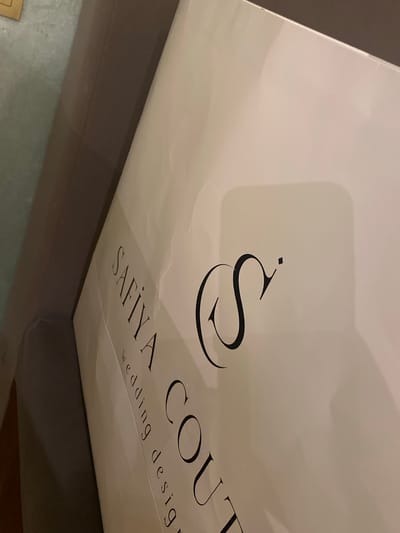 This media file captures a close-up, angled view of a white paper shopping bag with a prominent black logo and text. The bag appears to be made of a matte material and shows some minor creases and reflections from lighting.
The logo features a stylized, calligraphic "S" with a small diamond-shaped dot above it. Below the logo, the text "SAFIYA COUT" is visible in an elegant, serif font. Further down and slightly to the right, smaller text reads "wedding design". The overall presentation suggests a brand associated with high-end wedding attire.
The setting appears to be indoors, with the bag resting on a dark, textured surface, possibly a piece of furniture. The background is out of focus, but hints at a neutral or dimly lit environment. There are no people or specific activities depicted, the focus is solely on the product packaging. The lighting creates shadows and highlights on the bag's surface, emphasizing its shape and the texture of the paper. The image's composition and focus draw attention to the brand name and its association with "wedding design". Given the location context of Buynaksk, Russia, this bag likely belongs to a local boutique or is a purchase made within the region.