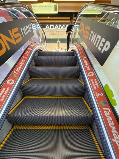 The image shows an upward-facing view of a shopping mall escalator. On the left and right sides of the escalator, large dark gray signs with orange and white text that reads "DNS ГИПЕР" are visible. The steps of the escalator are grey with yellow safety edges. A red band runs along the side of the escalator, featuring text in Russian, including "Галамарт" and "товары для дома," along with circular icons. A young man in a dark t-shirt and pants is standing on the escalator steps, looking at his phone. In the background, there is signage indicating directions and shop names. The ceiling of the mall, with its wooden slatted design and overhead lighting, is also visible. The overall environment suggests a commercial retail space in a modern shopping center, likely in Moskovskiy, Russia.