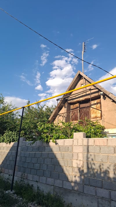 The image depicts an outdoor scene with a focus on an old building and its surroundings under a bright blue sky with scattered white clouds. The building, which appears to be a shed or small house, has a light brown, weathered facade and a peaked roof with exposed wooden beams. The windows are covered with rusty, corrugated metal sheets and decorative ironwork. Lush green vegetation, including leafy bushes and trees, grows around the building, partially obscuring it and adding a touch of nature to the otherwise dilapidated structure.
To the right of the building is a wall constructed from grey concrete blocks. The wall casts a distinct shadow, suggesting the sun is positioned in the sky, likely indicating it is daytime, possibly late afternoon or early morning given the angle of the shadows.
In the foreground and spanning across the image are several dark wires, one of which is very thin and runs diagonally. A bright yellow pole, possibly a conduit or support structure, also runs diagonally across the scene. A television antenna is visible mounted high on a wooden pole against the sky. The overall impression is one of a rural or semi-rural setting in Шұбарсу, Kazakhstan, characterized by older structures and overgrown greenery. There is no visible text in the image.