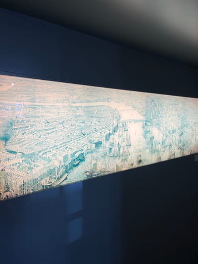 The image shows a framed illustration of the city of Vienna, Austria, located at the coordinates 48.194 latitude and 16.345 longitude. The illustration is mounted on a dark blue wall. The image is a bird's-eye view of the city, showing the city's buildings, streets, and waterways. The illustration is in grayscale and blue tones. The illustration appears to be lit from above, creating a bright spotlight over the image. The illustration is framed in a thin silver frame, and the image is in focus.