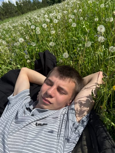 A young man is lying on a field of grass and dandelions. He is fair-skinned with short brown hair and closed eyes. He is wearing a striped gray and white short-sleeved collared shirt and resting on a black jacket. The location is a field in Дойная, Russia. The sun is shining, casting shadows of vegetation on the man's arm. The field extends into the distance with scattered trees on the horizon.