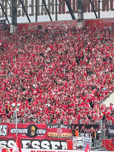 A large crowd of people wearing red and white clothing fills a stadium, likely at a sporting event. Several banners are visible, including one that reads "ESSEN" and others with various slogans and team names such as, "Northside Crew Essen" and "Crazy-Boy's". There is a sign near the top of the stadium reading "W2". The context suggests this is a soccer match in Essen, Germany, attended by many fans wearing the team colors, and many people are present to support their team.