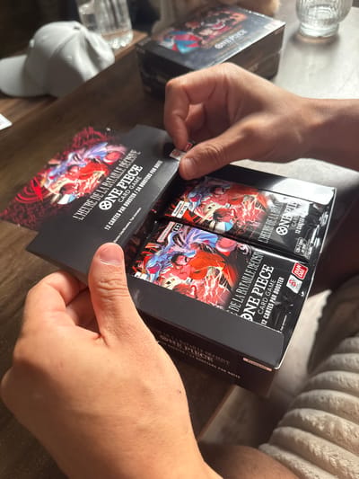 A close-up, high-angle interior shot depicts a person's hands in the process of opening a black cardboard display box of "ONE PIECE CARD GAME" booster packs. The hands, with visible thumbs and fingers, are light to medium-toned. The left hand stabilizes the bottom of the box, while the right hand lifts the top flap.

The box is predominantly black, with the product title "L'HEURE DE LA BATAILLE DÉCISIVE ONE PIECE CARD GAME" printed in white and red lettering on the top flap. Below this, it states "12 CARTES PAR BOOSTER / 24 BOOSTERS PAR BOITE" (12 cards per booster / 24 boosters per box), indicating it is a French language edition, further confirmed by a small "Français" label visible on the side of the box. Inside, multiple individual sealed booster packs are visible, stacked neatly. These packs are black with vibrant anime-style artwork featuring dynamic characters and energy effects, common to the One Piece franchise. The "BANDAI NAMCO" logo and a "6+" age rating are discernible on some of the booster packs.

The scene takes place on a dark brown, polished wooden or laminate table surface. In the background, to the left, a white baseball cap is visible, alongside a clear, textured glass, possibly containing water. Further back on the table, another identical or similar black box of One Piece cards is present. The lower right and bottom center of the frame reveal a light-colored, textured fabric, likely a blanket or cushion. There are no identifiable visual cues or landmarks within the image to determine the specific city of Wattrelos, France, or any other precise geographical location.