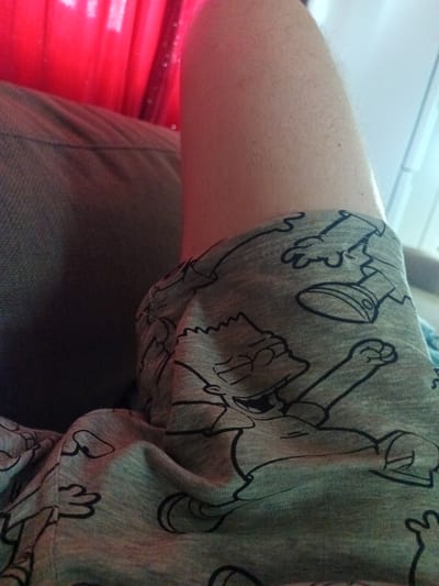 The image is a close-up shot of a person's leg wearing grey pajama bottoms with a cartoon print. The print appears to be of Bart Simpson and other characters from The Simpsons. The leg is resting on a dark-colored surface, possibly a couch or chair, and a red, translucent fabric, likely a curtain, is visible in the background. The lighting suggests an indoor setting. The image does not contain any information about the location being Risøyhamn, Norway.