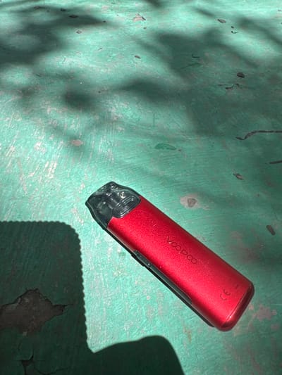 A red, metallic vape pen with a clear plastic mouthpiece rests on a cracked, teal-painted surface. The surface is uneven and shows signs of wear and tear, with areas of peeling paint and exposed concrete. Shadows from overhead foliage create dappled patterns on the ground. The vape pen displays the "VooPoo" logo in white lettering and has a small "CE" marking near its base. The image appears to be taken outdoors on a sunny day, likely in an urban environment, though specific details placing it in Termirtaw city administration, Kazakhstan are not discernible from the visual information provided.