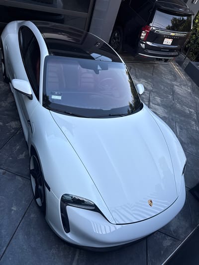 A high-angle shot depicts a white Porsche Taycan parked on a grey, patterned concrete driveway in what appears to be a residential area. The vehicle is positioned near a dark SUV, which shows a California license plate reading "9TIW264." The scene implies a location in West Hollywood, United States of America, given the California license plate. The light suggests daytime conditions.