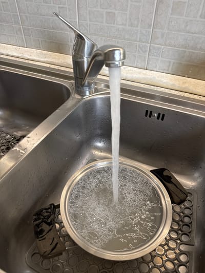 The image shows a stainless steel kitchen sink in Aprilia, Italy. A stream of water flows from a chrome faucet into a metal pot filled with soapy water. The pot sits on a round, perforated silicone mat. The sink has a rectangular overflow drain. The counter beside the sink is tiled.