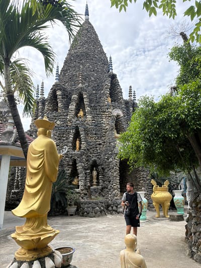 This image depicts an outdoor scene at a unique religious or spiritual complex, likely in Cam Ranh, Vietnam, characterized by its distinctive stone architecture and numerous statues.
The central subject is a towering, conical structure, possibly a stupa or pagoda, constructed from rough, dark gray stones or rocks. This multi-tiered edifice features numerous small, golden figures or statues placed within niches across its surface. Intricate spires with stacked, ring-like elements adorn various points of the structure, extending towards the sky.
In the foreground, a large golden statue, possibly of a monk or deity, is seen from behind, wearing a hat or headpiece and flowing robes. Another golden statue's head and torso are visible further right, next to a paved pathway. In the background, two large, decorative urns or vases stand on pedestals, one golden and the other light blue/green with painted patterns.
A male visitor is present on the paved ground to the right of the main stone structure. He has short hair and a beard, and is dressed in a black vest over a lighter shirt, black shorts, and carries a black bag. He appears to be observing the site.
The scene is framed by vibrant green foliage, including palm fronds in the lower left and lush trees and bushes growing around and above the stone structure. The sky is bright and partly cloudy, indicating daytime. The atmosphere is calm and contemplative, reflecting the sacred nature of the site. There is no visible text in the image.