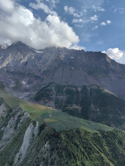 The image depicts a vast mountainous landscape with a prominent ridge in the foreground. The ridge, covered in a mix of green vegetation and rocky outcrops, slopes downwards and to the left. Patches of dark green trees are scattered across the slopes. Further back, a wider, gentler slope of green terrain is visible, with some rectangular fields, suggesting agricultural activity. In the far distance, massive, dark-colored mountains rise steeply, their peaks partially obscured by thick, white clouds. The sky is blue with scattered white clouds. A narrow, gray path or stream bed cuts through a valley between the foreground ridge and the distant mountains. The overall scene is one of natural grandeur, with a sense of remoteness. The image is a landscape photograph, likely taken from an elevated position, offering a panoramic view of the mountainous terrain. The specific location in Tiling, India, is suggested by the context but not visually evident within the image itself. There are no people or animals visible in the frame.