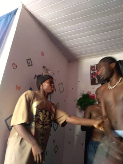 In Kassa Market, Nigeria, two young men are interacting in an indoor space. The man on the left, wearing a tan t-shirt with a graphic print and a black beanie, is turned to his right, appearing to speak or gesture. To his right, a man with braided hair, wearing a necklace and blue shorts, is mostly out of focus, facing left. The background features a wall decorated with various small, colorful shapes, including hearts and butterflies, and some framed pictures. A shelf with a green plant and pink flowers is visible. The ceiling is made of white, horizontal panels. The lighting suggests it is daytime, with light coming from the left.