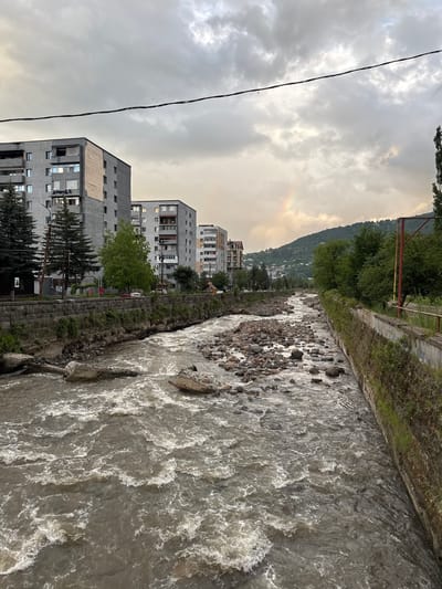 The image captures a view of the Dilijan river in Dilijan, Armenia, under a cloudy sky with a faint rainbow visible in the distance. The river flows through the town, with concrete banks and a stony riverbed. On the left side of the river, there are several multi-story residential buildings and evergreen trees. On the right side, the riverbank is lined with vegetation and a concrete wall, with a red and yellow pole visible in the background. The water appears murky and turbulent, with rocks and debris scattered throughout its course. The overall atmosphere is overcast, suggesting recent or impending rain.