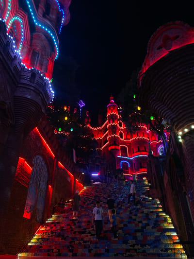 The image shows a vibrant night scene at the "Forbidden City" tourist attraction in Baqiao, China. The main subject is a grand, castle-like structure adorned with numerous colorful lights, primarily red and blue, outlining its architectural features. A long, wide staircase, covered in a mosaic of colorful tiles, leads up towards the illuminated structure. Several individuals are visible on the stairs, walking up and down, some in groups and others alone. The overall impression is one of a festive and visually striking nighttime tourist destination, with the lights and colorful stairs creating a magical atmosphere. The environment appears to be an outdoor public space, likely a park or cultural attraction, with trees visible in the background, partially obscured by the lights and the structure. The darkness of the night sky contrasts sharply with the bright illumination of the attraction, emphasizing its visual impact.