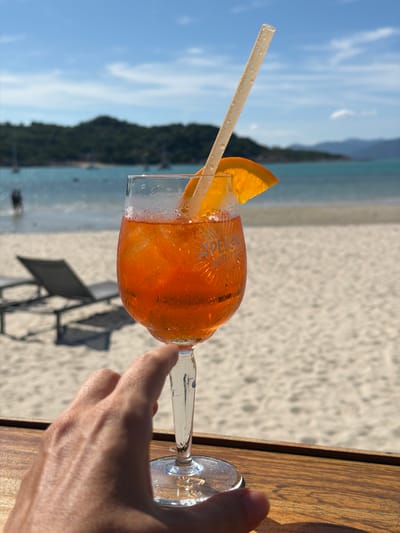 A hand with tanned skin and visible veins is reaching towards a stemmed glass filled with an orange aperitif. The glass is sweating from condensation, and a thin, light-colored straw extends from the drink, topped with a slice of orange. The glass has "APEROL SPRITZ" etched into it. The drink and glass are positioned on a wooden surface. In the background, a sandy beach stretches towards a calm, turquoise ocean under a clear blue sky with scattered white clouds. Two empty lounge chairs are visible on the sand, and in the distance, lush green hills rise from the coastline. The overall scene evokes a relaxed vacation atmosphere in a tropical location, possibly Choeng Mon, Thailand, given the sandy beach, clear water, and lush vegetation.