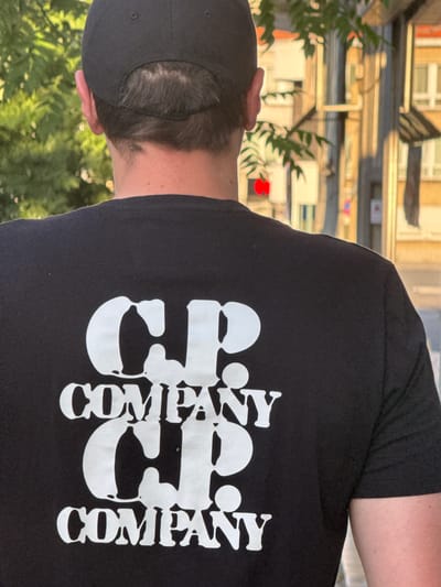 A man stands with his back to the camera, wearing a black t-shirt with white "C.P. COMPANY" lettering on the back. He is also wearing a black baseball cap. The background shows a blurred urban scene with buildings and trees. The image appears to be taken outdoors during the daytime. The man is positioned centrally in the frame, and the focus is on the t-shirt graphic. The lighting suggests it might be late afternoon or early evening, with warm, golden light illuminating the scene.