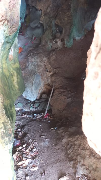 The image depicts the interior of a cave, likely located in Thap Put, Thailand. The cave's roof is visible at the top of the frame, composed of rough, light-colored rock. Inside, the walls are uneven, with brown and gray hues and a mix of smooth and jagged textures.

On the left, there's a deposit of green-tinted rock. At the bottom center, a ledge of green rock is visible.

A wooden beam is lodged horizontally between two walls, and some red cloth is nearby.

On the right, the cave floor is covered with fallen leaves and loose soil.