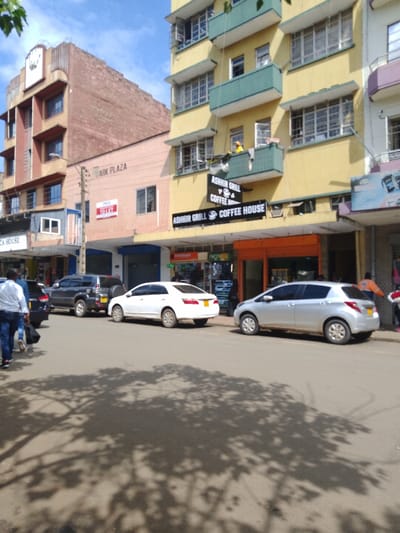 The image shows a street scene in Athi River, Kenya, at the coordinates -1.453 latitude and 37.007 longitude. The street is lined with buildings, including a tall building with the sign "Arik Plaza" on the side. There is a coffee shop with the sign "Ashir Grill Coffee House" on the front. There are four cars parked on the street: a black SUV, a silver hatchback, a white sedan, and a black sedan. There are several people walking in the street. The sky is partly cloudy and there is a large shadow of a tree on the street. The street is paved and appears to be relatively quiet.