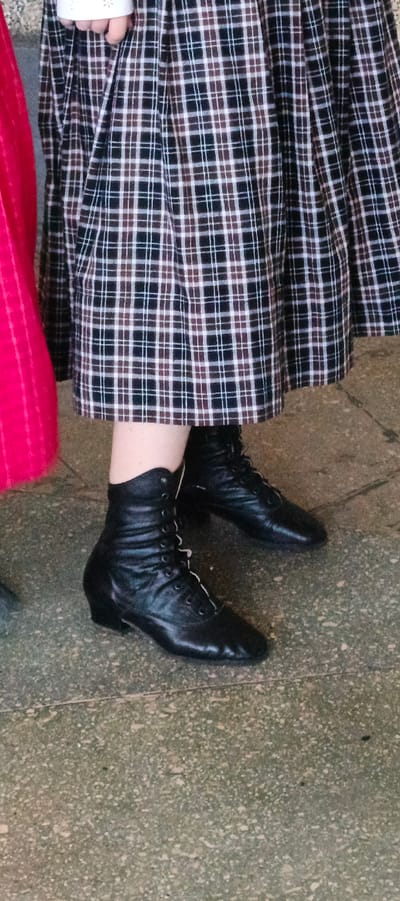 The image features a portion of a person wearing a pleated, plaid skirt in shades of black, brown, and white. They are also wearing black, lace-up ankle boots. A small section of a red garment is visible to the left of the person. The ground appears to be a speckled, light gray surface. The photograph was taken in the city of Кемуль, Russia.