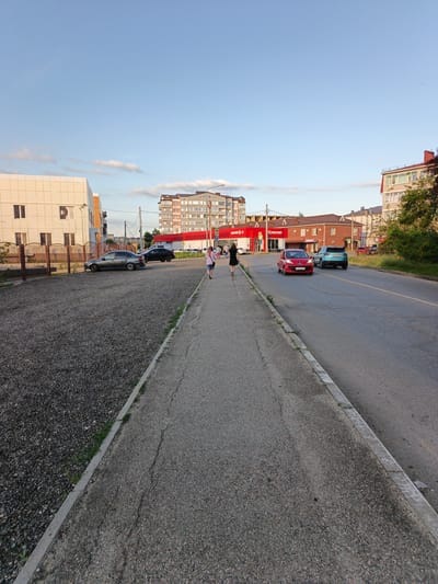 The image depicts a street scene in Goryachiy Klyuch, Russia, with a clear blue sky and scattered clouds overhead. In the foreground, a paved sidewalk runs parallel to a road. To the left of the sidewalk is a gravel parking area with a dark gray sedan and a black SUV parked. A low metal fence separates the gravel area from a modern, light-colored building with dark-trimmed windows.
The road itself has two lanes and is occupied by a red hatchback and a turquoise SUV. Two pedestrians, a woman in a black dress and another person in a light-colored top and dark pants, are walking away from the camera on the sidewalk. In the background, several multi-story buildings are visible, including a prominent red building with white lettering that appears to be a store. The overall impression is of a suburban or small-town street in the late afternoon or early evening.